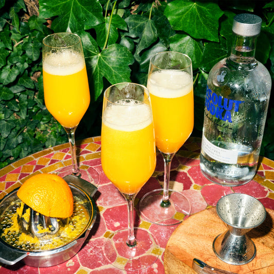 3 orange mimosas shown next to a vodka bottle on a mosaic table in front of green leaves
