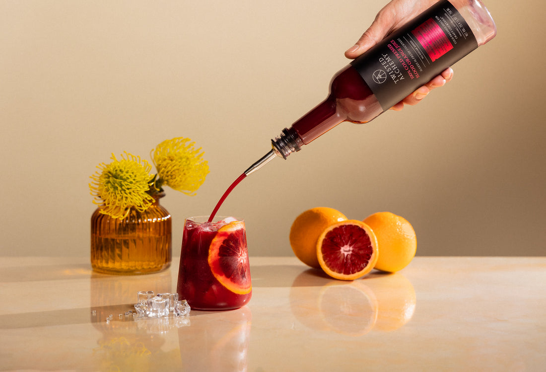 Hand pouring blood orange juice into a stemless glass garnished with a blood orange wheel, with flowers and blood oranges in the background