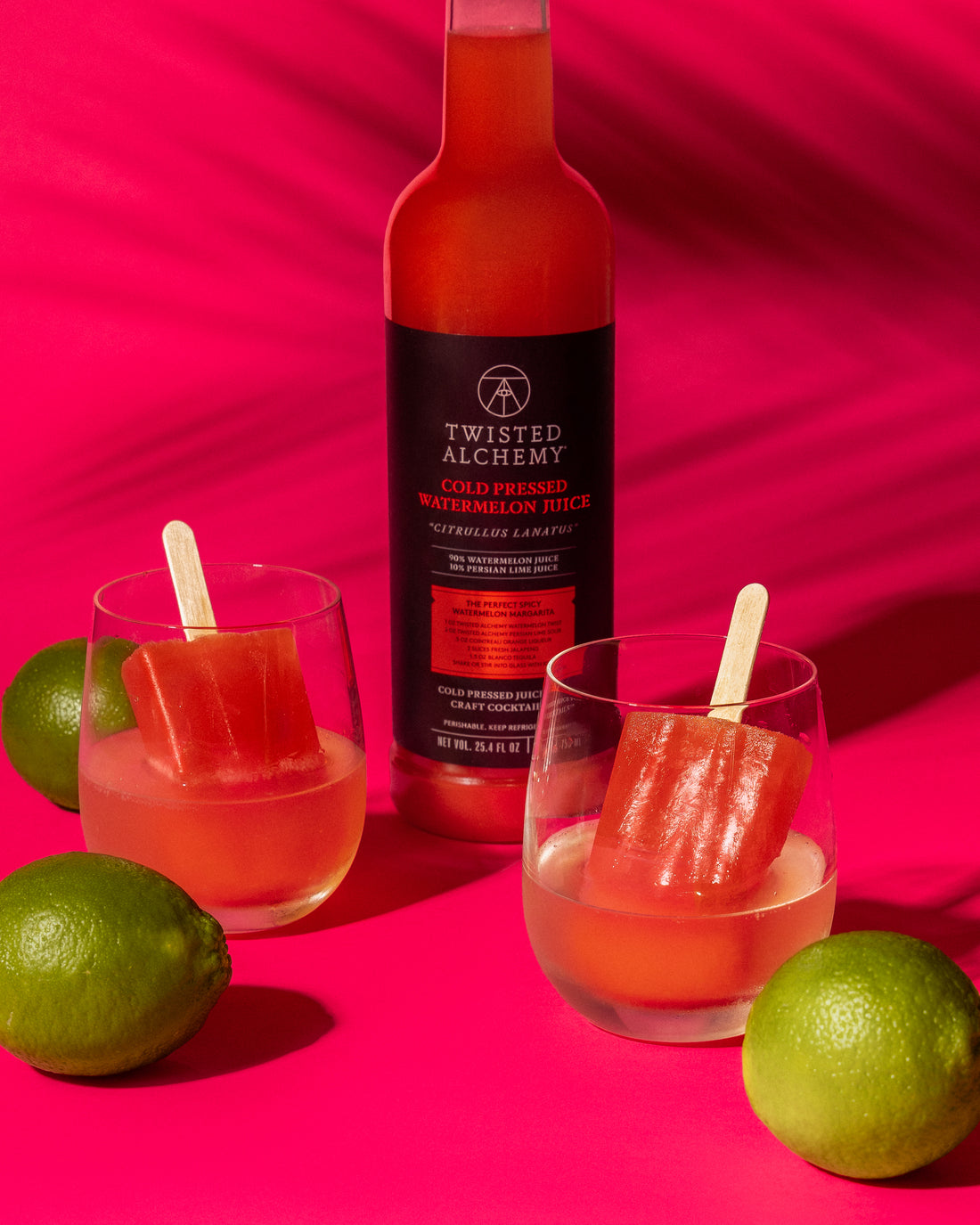 Two popsicles in glasses in front of a bottle of Twisted Alchemy watermelon juice, surrounded by limes