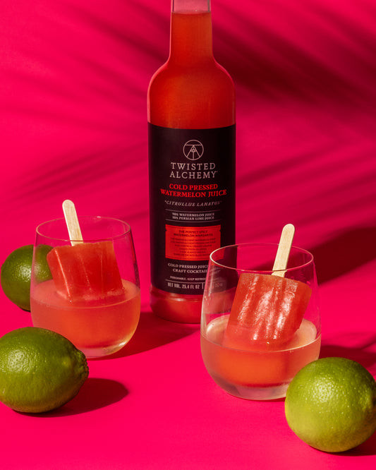 Two watermelon popsicles in stemless glasses surrounded by whole limes and a bottle of watermelon juice against a pink background