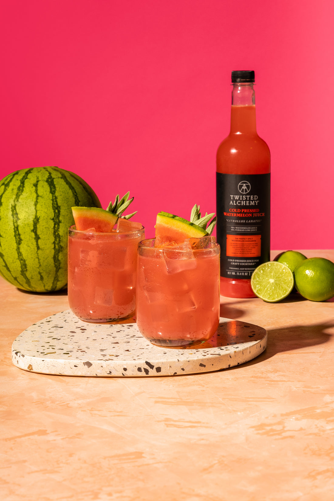 Two cocktails with watermelon slice garnishes sit in front of a bottle of watermelon juice next to three limes and a whole watermelon.