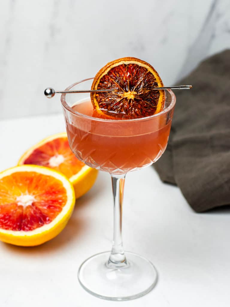 Blood orange cocktail shown in glass on white table with dried blood orange on a toothpick across the drink