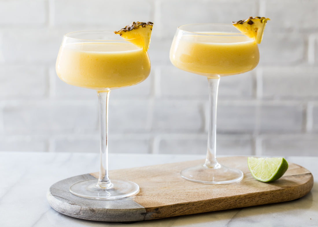 2 pineapple frozen cocktails in round glasses with stems on a wooden circle against a white brick wall. There is a lime slice next to the glass.