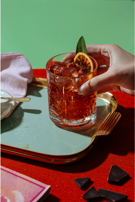 Hand holding a red cocktail in a glass with a dried citrus garnish and a leaf garnish on a blue and gold trey against a red and green background.