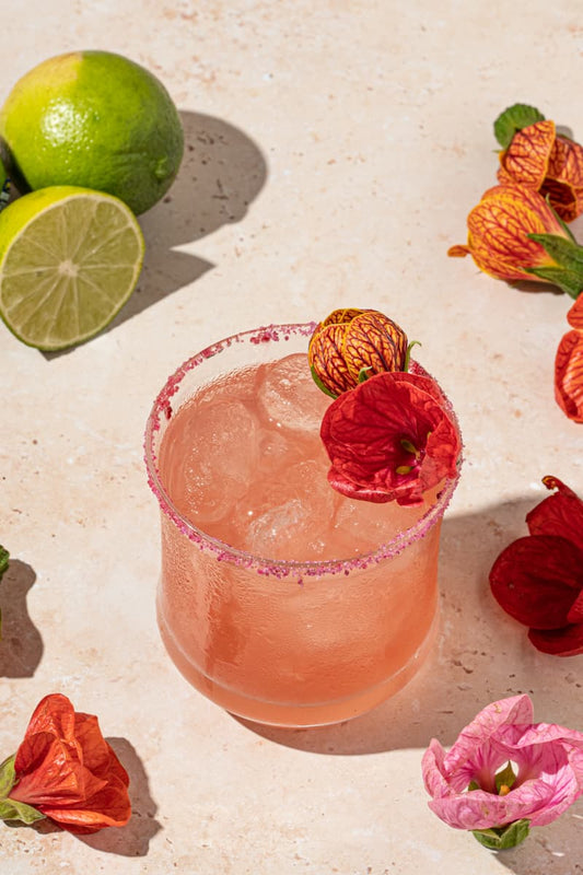 Pink colored cocktail inside a glass with ice, a purple salt rim, and a hibiscus flower garnish. Surrounded by a lime half, and more hibiscus flowers.