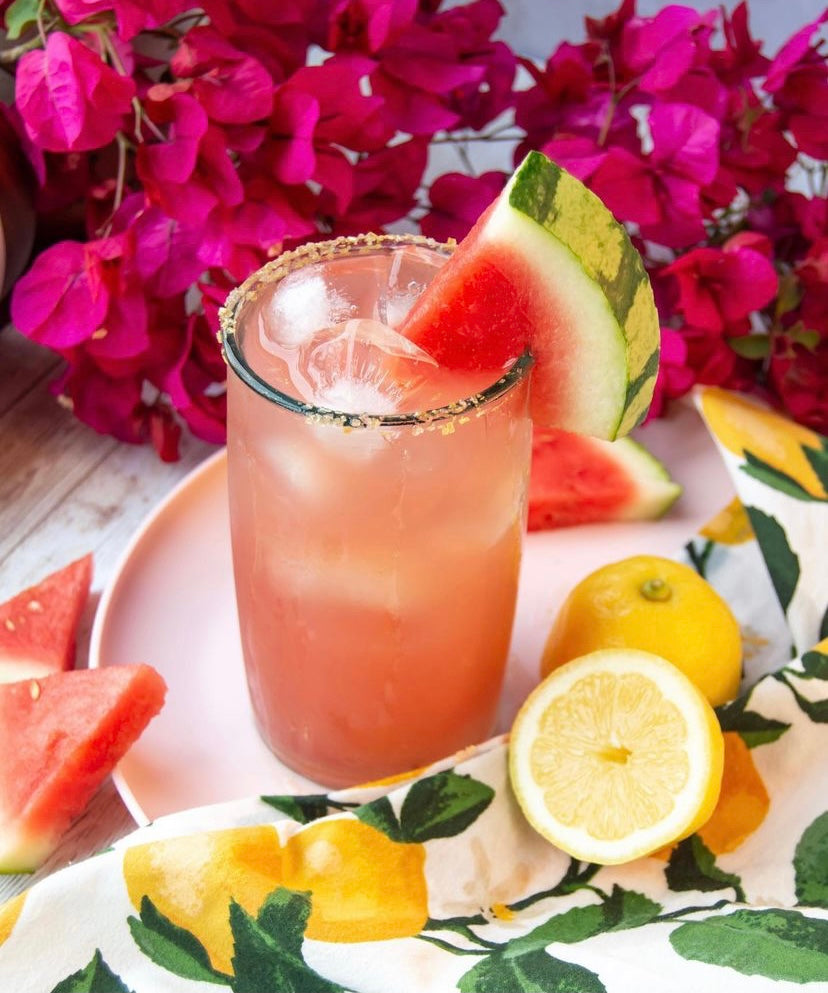 Watermelon margarita pictured on white plate with fresh lemon slices and watermelon slice. There are pink flowers in the background