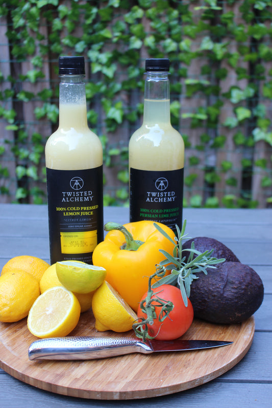 Two bottles of juice sit on a cutting board with limes, lemons, avocados, peppers, and tomatos.