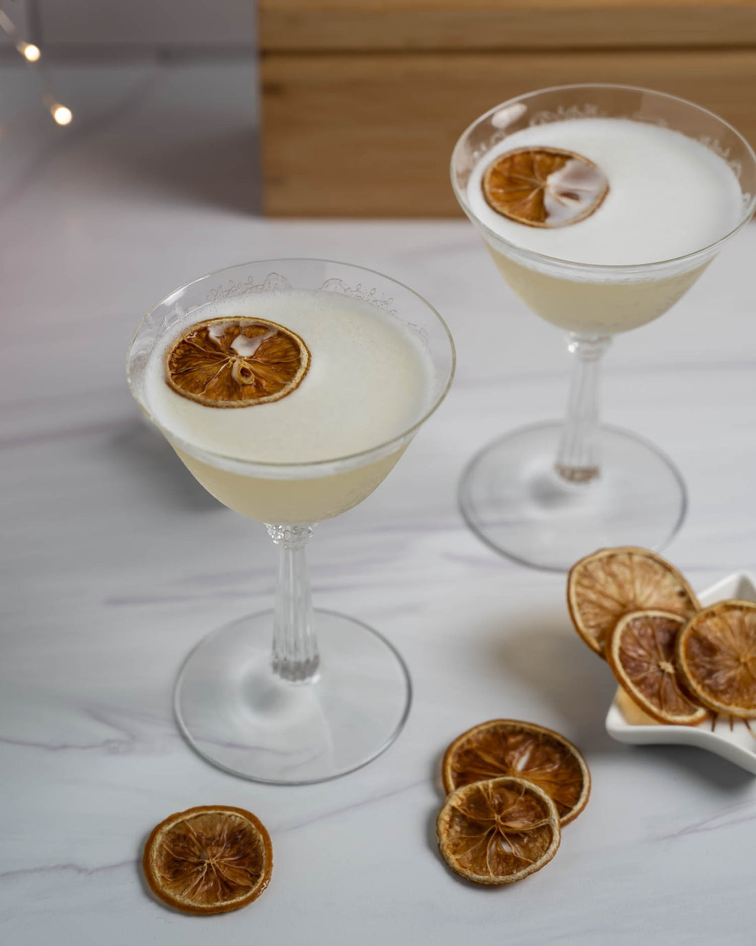 Two lemon cocktails with foam on top, topped with dried citrus wheel on marble tabletop with wood behind it
