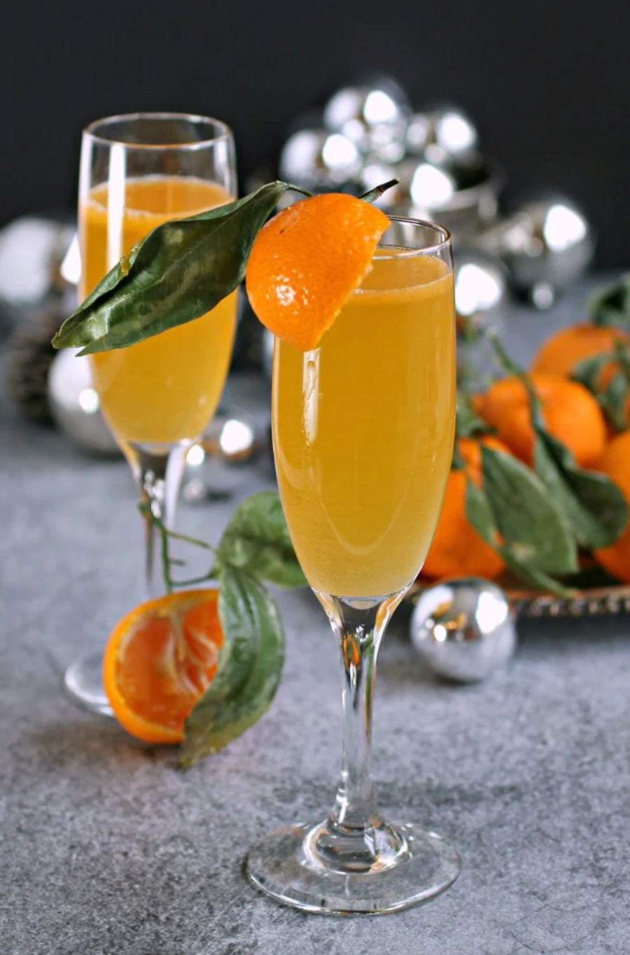 Two mimosas in champagne glasses with orange wedges. Oranges with their leaves left on sit in the background.