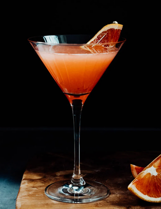 Blood orange martini with a blood orange slice on a wooden cutting board against a black background