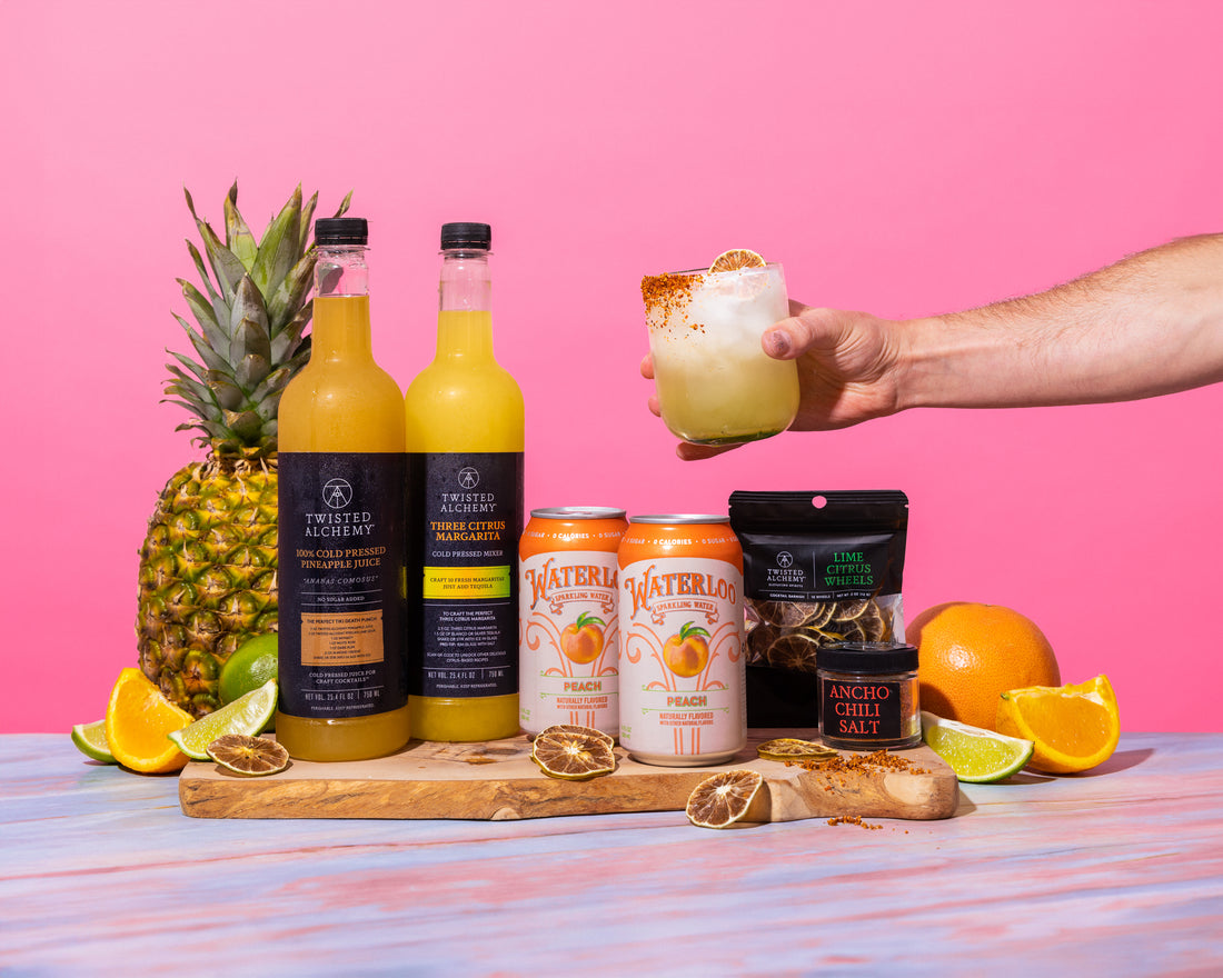 A hand holding a cocktail over a kit including two bottles of Twisted Alchemy juice, two bottles of Waterloo soda, and garnishes on a cutting board surrounded by pineapples, oranges, and limes.