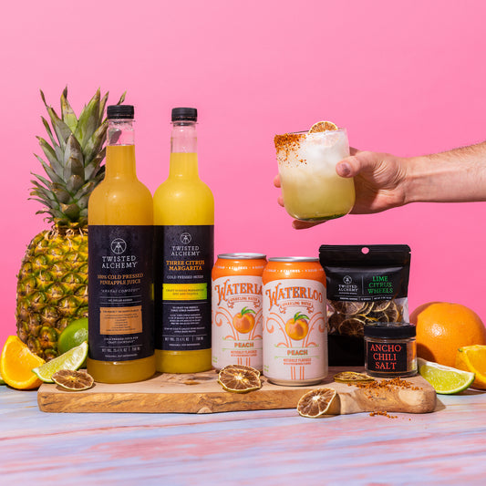 Hand holding a glass garnished with a lime wheel and red chili salt over a cutting board with two juices, two cans of soda, dried lime wheels, a chili container, and cut citrus fruits against a pink background