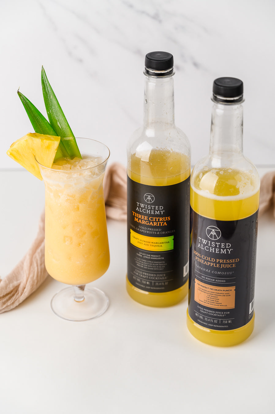 Two bottles of twisted alchemy juice next to a rum cocktail with a pineapple wedge and pineapple leaves