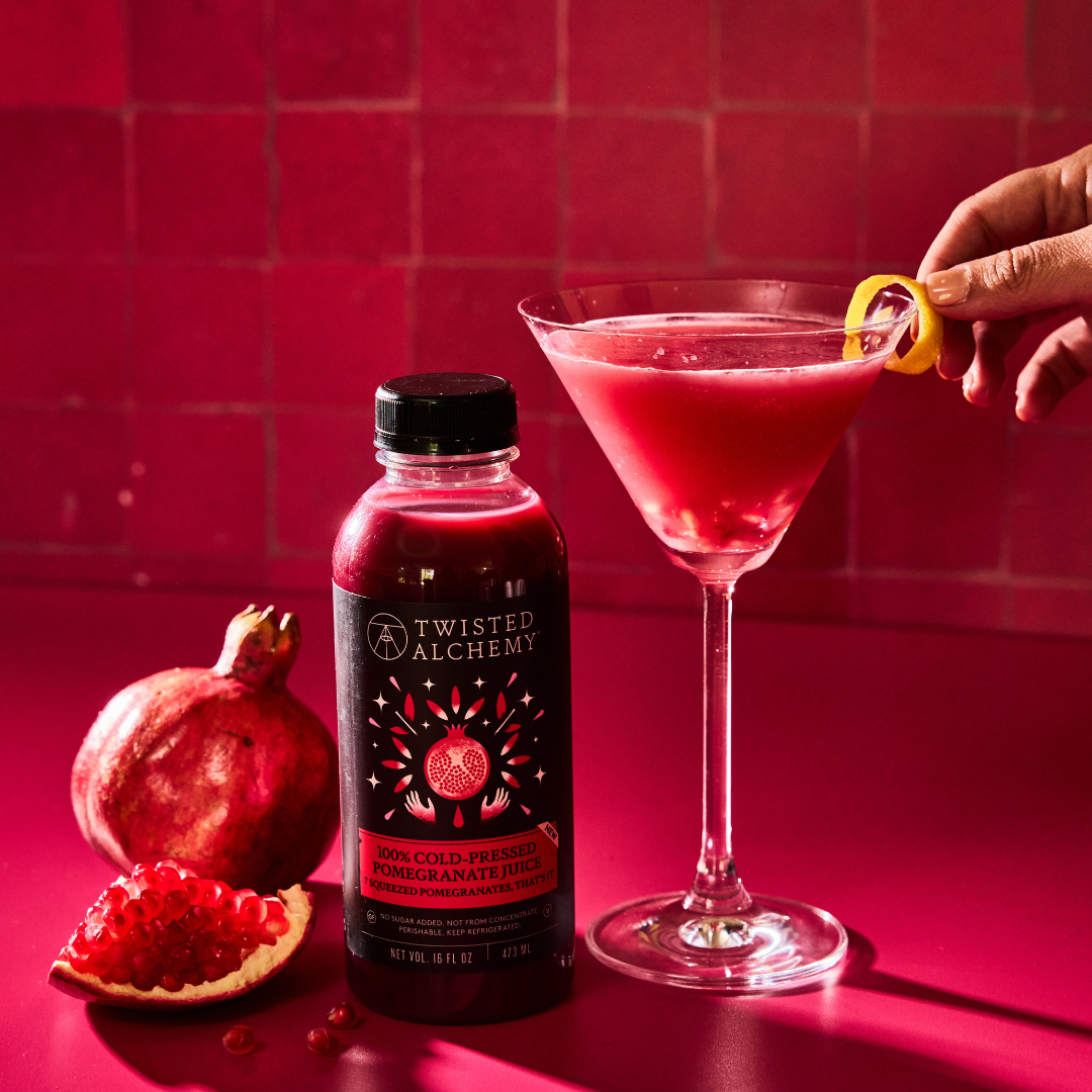 Twisted Alchemy Pomegranate juice next to a red pomegranate martini styled with lemon garnish