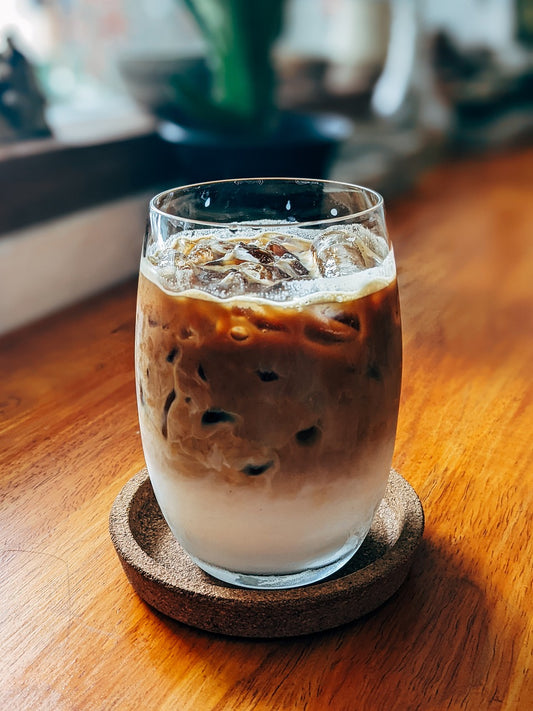 Iced coffee in a stemless glass on a cork coaster against a wooden table. 