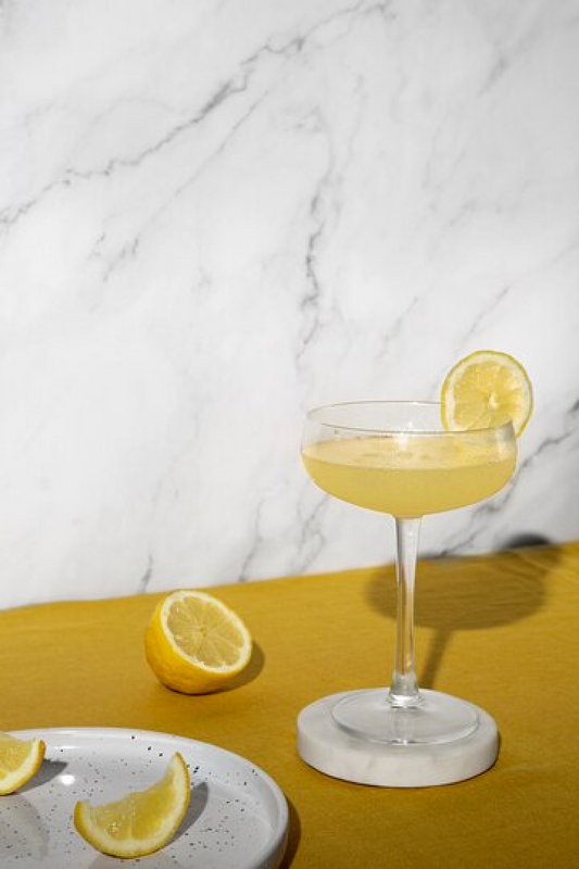 A coupe glass on a marble coaster with a yellow cocktail in it and a lemon slice garnish. Surrounded by a lemon half, and two lemon wedges on a marble plate.
