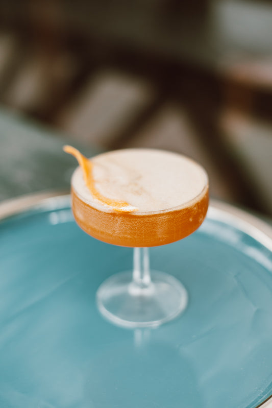 A whiskey cocktail in a coupe glass topped with eggwhite foam and an orange peel garnish, on top of a blue surface.