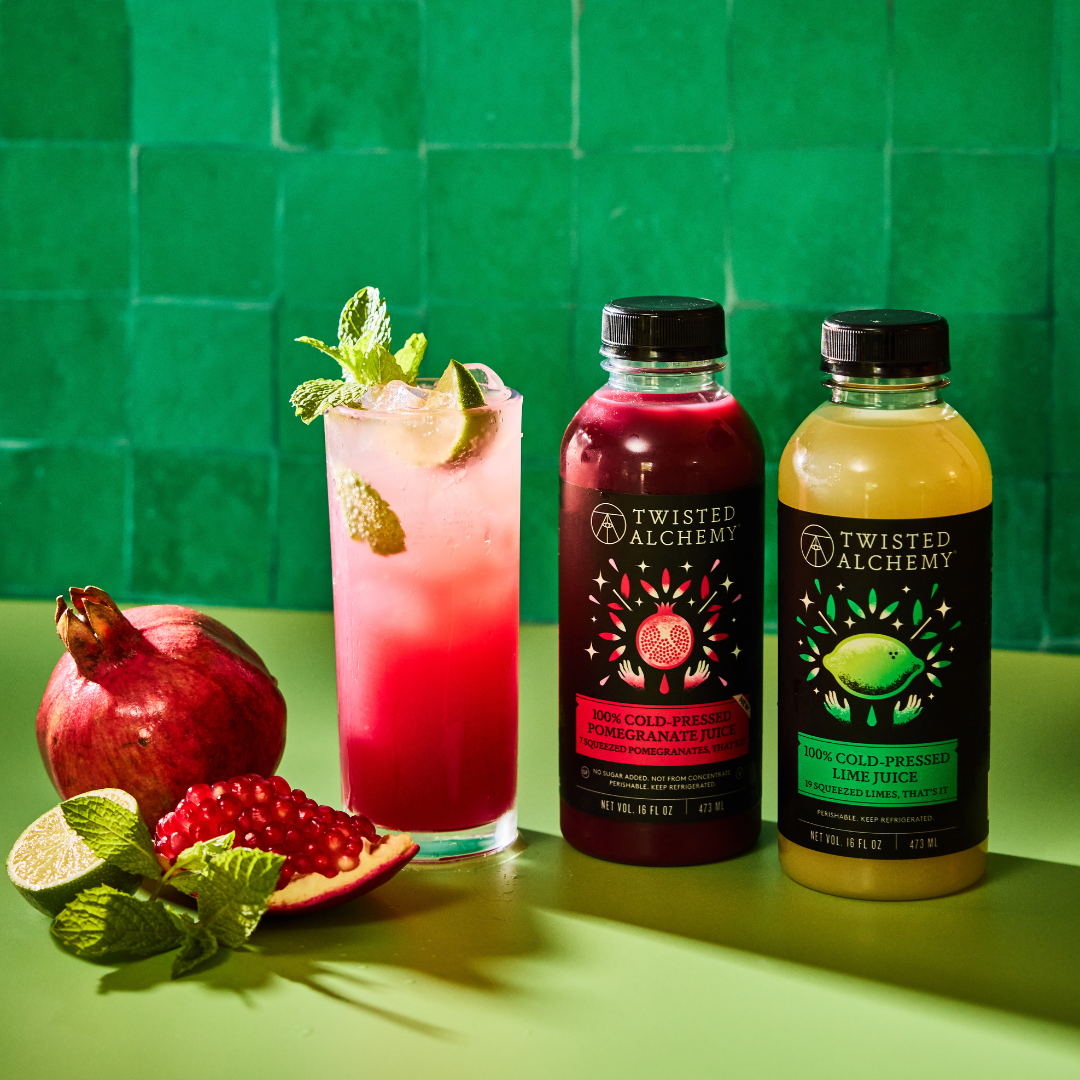 Two Twisted Alchemy juice bottles next to a red cocktail and pomegranate