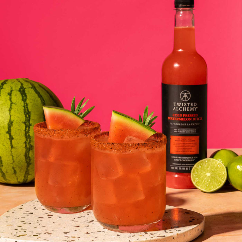 Two glasses of red cocktail with watermelon slices, a bottle of Twisted Alchemy juice, and a watermelon on a pink background.