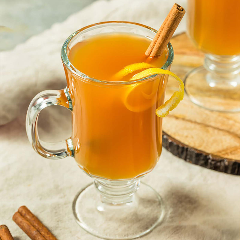 Glass mug of hot apple cider with a cinnamon stick and lemon peel on a wooden surface.