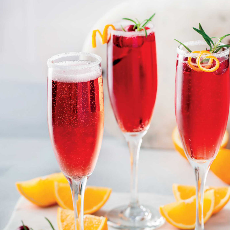 Three champagne flutes filled with a red sparkling beverage, garnished with orange slices and herbs, on a light background.