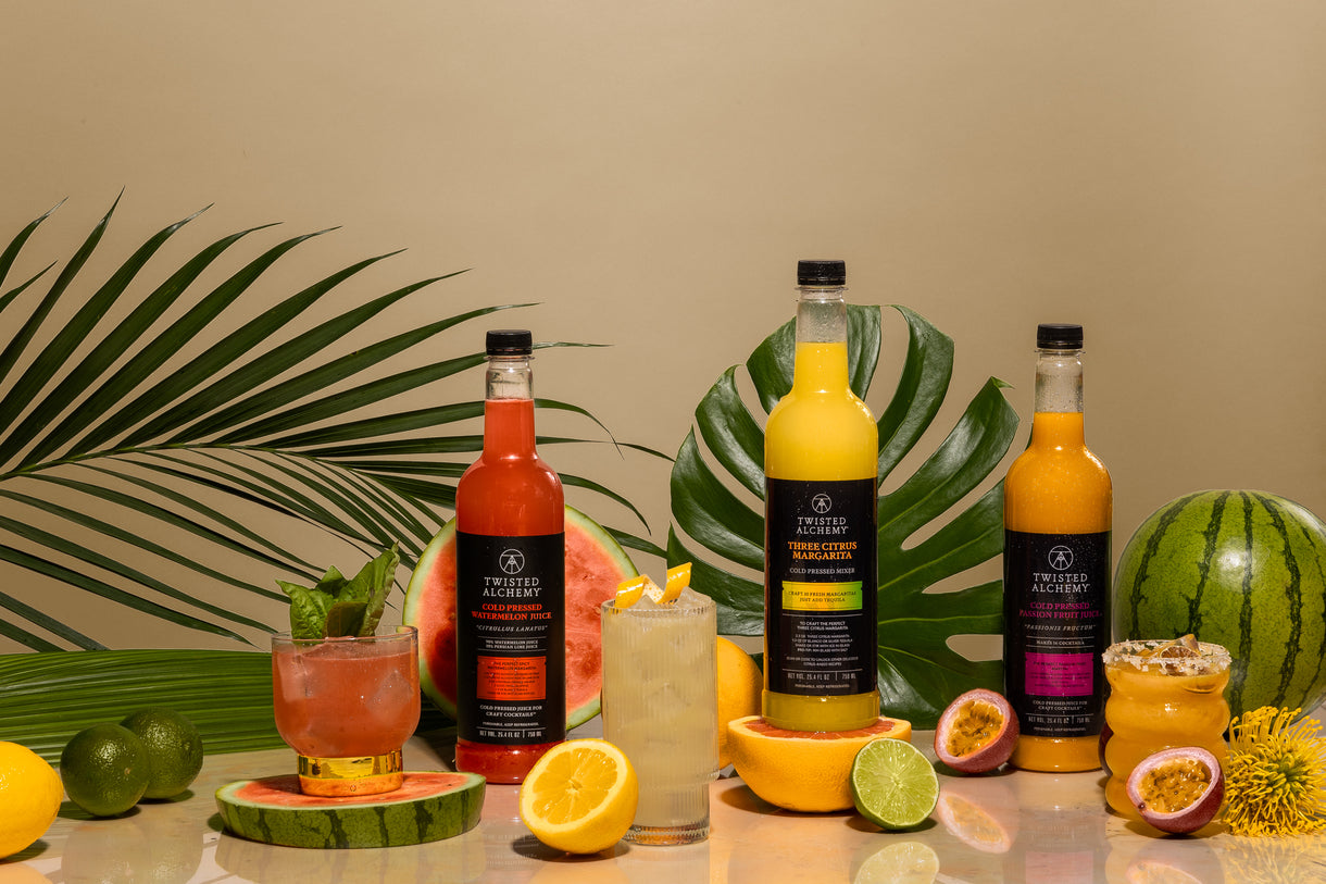 Three bottles of various Twisted Alchemy cocktail juices surrounded by fruits and leaves on a tan background