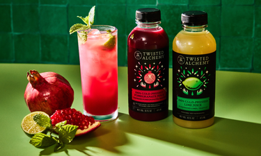 Two bottles of Twisted Alchemy juice with a cocktail and pomegranate fruit on a green background.