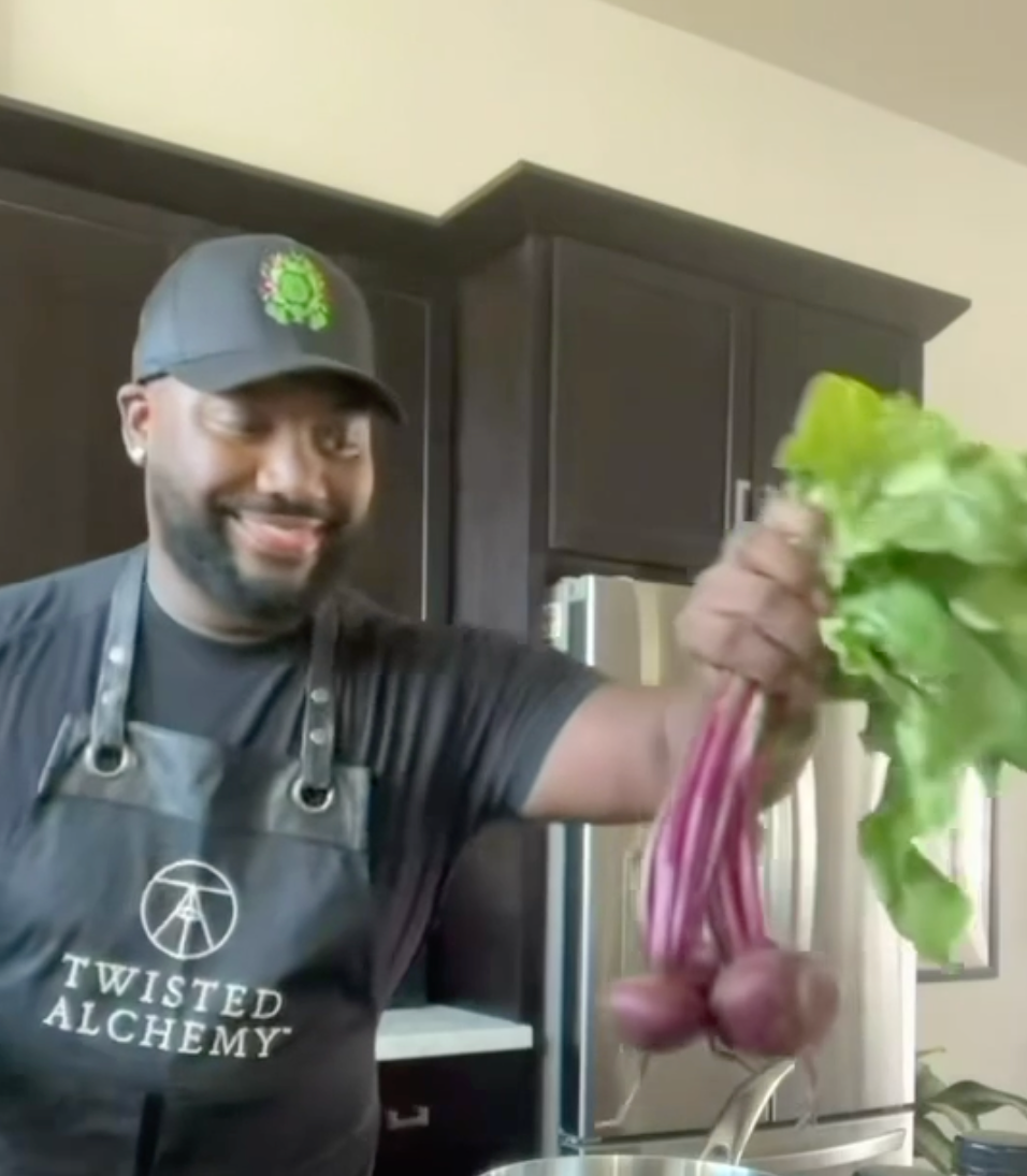 Person in a kitchen holding beets and a bottle, wearing a 'Twisted Alchemy' apron.