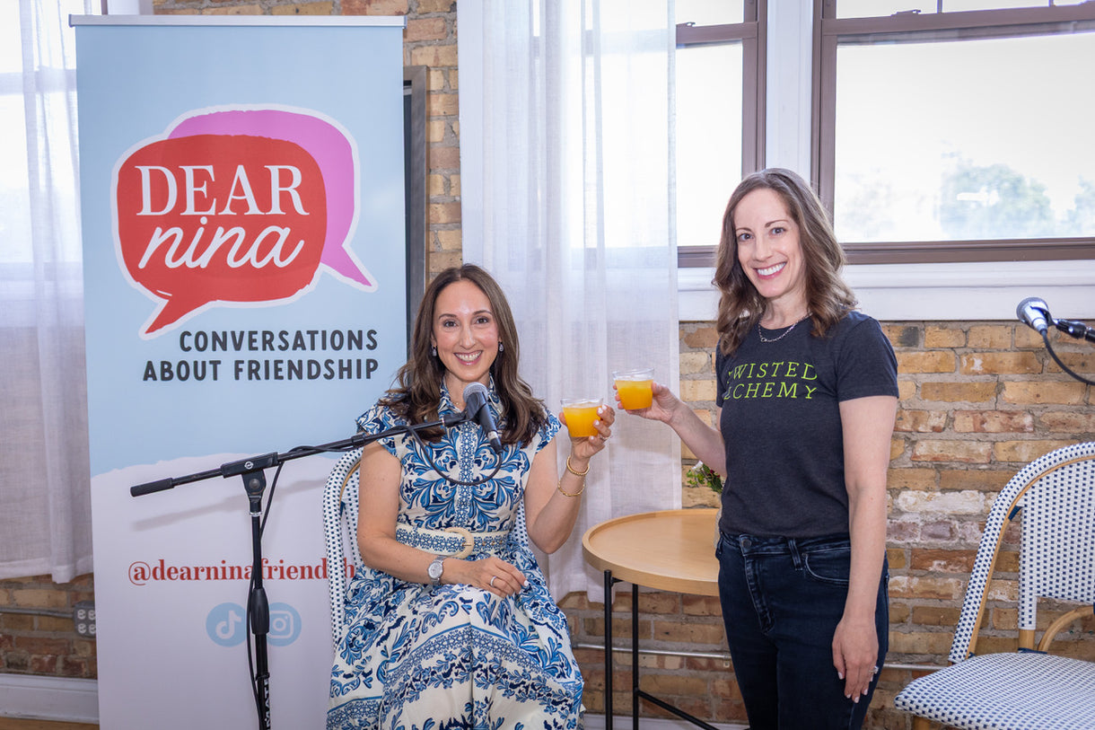 Two women standing next to a 'Dear Nina' banner holding Twisted Alchemy cocktails.