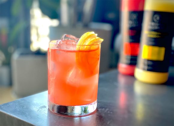 Bright orange cocktail in front of a pair of Twisted Alchemy bottles on a counter